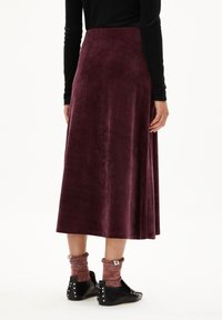 Jupe maxi en velours bordeaux avec une texture lisse, dotée d'une taille haute et d'une silhouette fluide. Assortie avec des chaussures noires et des chaussettes à motifs.