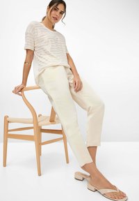 Lichte, gestreepte crèmekleurige gebreide top gecombineerd met beige op maat gemaakte broek en nude blokhak sandalen. Houten stoel met een geweven zitting.
