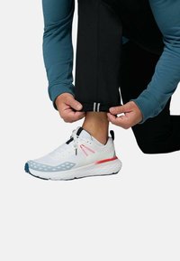 Personne ajustant le revers d'un pantalon noir en portant une chaussure de sport blanche avec des accents rouges et bleus sur un fond blanc.