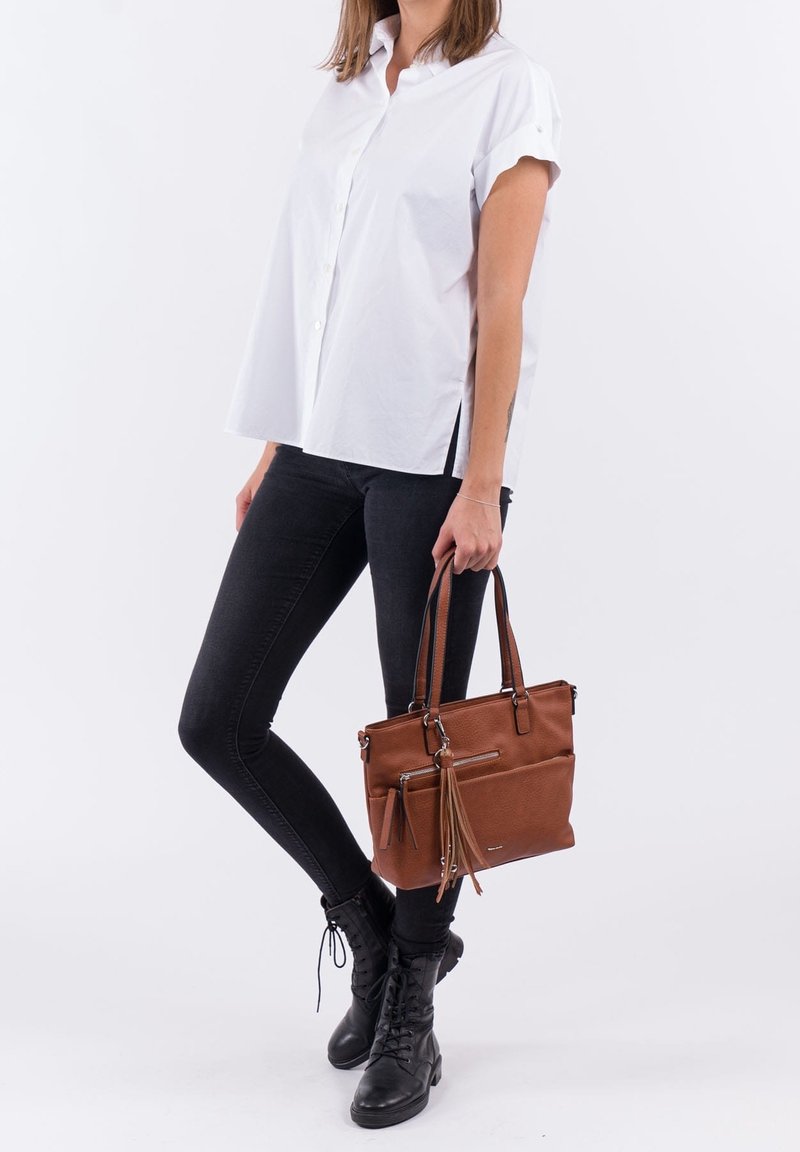 Femme portant une chemise blanche à manches courtes et un jean noir, tenant un sac à main en cuir marron avec un détail à franges, debout sur un fond blanc.