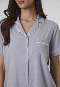 Grå kortärmad pyjamasöverdel med ribbad textur, vit kant, knäppning framtill och en enda ficka på vänster bröst.