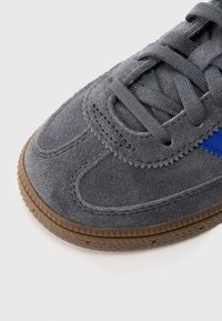 Pelēks zamša sporta kurpes ar ziliem akcentiem, raksturīgas ar fakturētu virsmu, apaļu purngalu un gumijas zoli. Šņores ir tumši pelēkā krāsā.