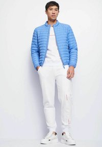 Giacca trapuntata blu chiaro con colletto alto, indossata sopra una camicia bianca e pantaloni della tuta bianchi con accenti rossi, abbinata a sneaker bianche.