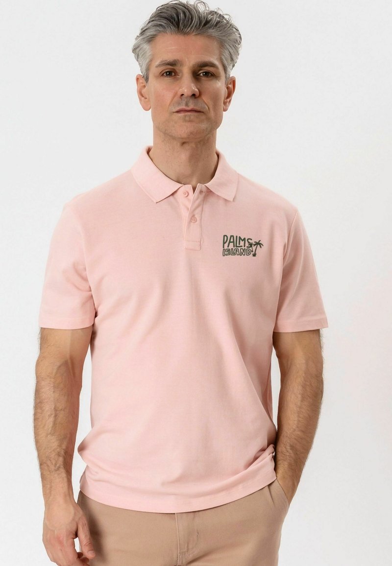 Homme portant un polo rose clair avec le texte « PALMS ISLAND » et un motif de palmier sur la poitrine, pantalon beige, debout devant un fond uni.
