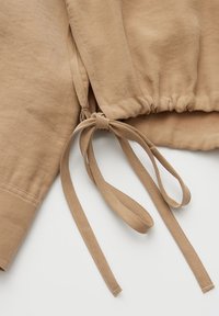 Hellbrauner Stoffärmel und Taillendetail mit Knotenschleife und elastischem Raffabschluss auf hellem Hintergrund.