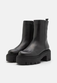 Sorte ankle boots i svart skinn med en glatt overflate, blokkhelt og tykk såle med et taggete mønster. Glidelåslukking på siden.