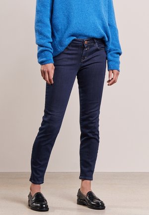 Person iført mørkeblå skinny jeans, en lys blå sweater og sorte læder loafers, der står på en lys gulv mod en ensfarvet baggrund.