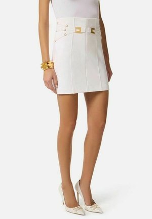 Donna con minigonna bianca con dettagli a fibbia dorata, tacchi alti bianchi e braccialetti dorati spessi al polso sinistro.