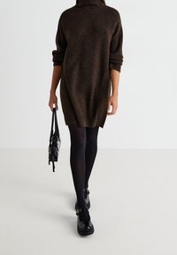 Abito in maglia marrone con collo alto, maniche oversize e vestibilità comoda. Abbinato a collant neri e scarpe nere lucide.