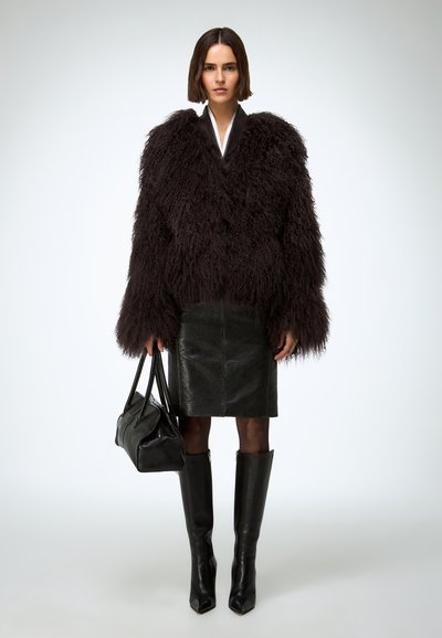 Czarna futrzana kurtka z teksturowanymi rękawami, zestawiona z dopasowaną czarną skórzaną spódnicą. Modelka trzyma czarną torebkę i nosi wysokie czarne kozaki.