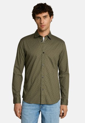 Mann mit dunkel lockigem Haar, der ein olivgrünes, langärmliges Hemd mit kleinen Punktmustern und hellblaue Jeans trägt.