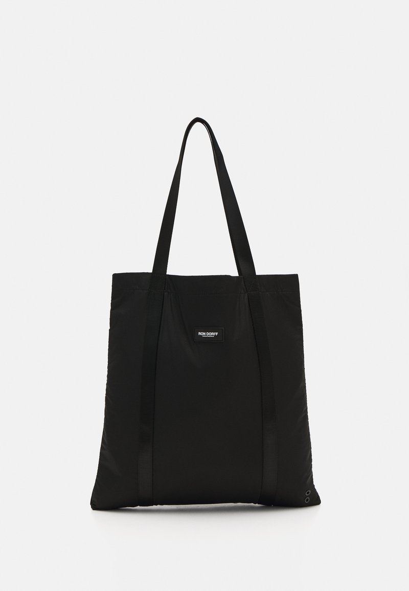 Svart tote bag laget av slitesterkt stoff, med to lange stropper, rektangulær form og en merkeetikett på fronten.