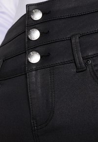 Pantalon en denim noir avec trois boutons argentés brillants à la taille, présentant une texture lisse et une coupe ajustée.