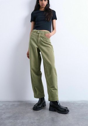 Jeune femme debout contre un mur blanc, portant un haut noir à manches courtes, un pantalon large en velours côtelé vert olive et des bottes à plateforme noires.