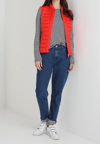 Röd kviltad utan krage väst över en svart- och vitrandig långärmad skjorta, i kombination med högmidjade blå jeans och vita sneakers.