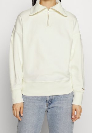 Frau trägt einen cremefarbenen Pullover mit Viertel-Reißverschlusskragen und hellblaue Jeans, steht mit locker an den Seiten hängenden Händen.