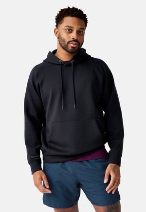 Homme debout portant un sweat à capuche noir, un short bleu marine, un t-shirt violet en dessous, avec un tatouage sur la main gauche et une boucle d'oreille à l'oreille gauche.
