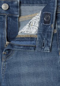 Tmavomodré rifle z denimu s poklopcom na zips, zlatým prešívaním a značkovým gombíkom. Interiér ukazuje svetlú vzorovanú látku.