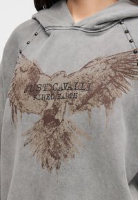 Γκρι φούτερ με κουκούλα, με ξεθωριασμένο γραφικό αετού, που αναγράφει "Just Cavalli" και "Fired Earth". Διακοσμημένο με μεταλλικές τρουκς στους ώμους.