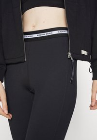 Leggings nere a coste con un alto punto vita, caratterizzati da un logo bianco. Il tessuto ha una consistenza liscia, accentuata da una giacca con zip.