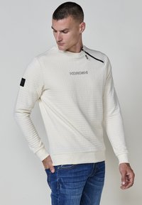 Sudadera color crema con un patrón texturizado acolchado. Presenta un cuello con cremallera y un parche lateral. Puños y dobladillo ajustados.