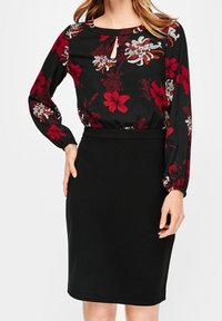 Femme portant une jupe crayon noire et un chemisier noir à manches longues avec un motif floral rouge et blanc et un décolleté en trou de serrure.