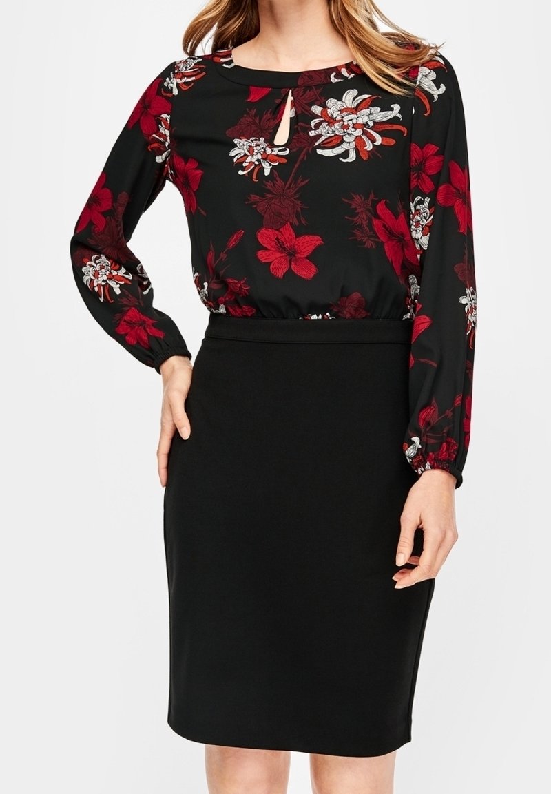 Femme portant une jupe crayon noire et un chemisier noir à manches longues avec un motif floral rouge et blanc et un décolleté en trou de serrure.