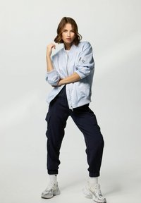 Light blue bomber jacket with ribbed cuffs, worn over a white top, paired with navy cargo pants and white sneakers, featuring a zip and pocket detail.