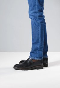 Chaussures en cuir noir avec une finition texturée, des panneaux latéraux élastiques et une semelle en caoutchouc épaisse. Portées avec un jean en denim bleu.