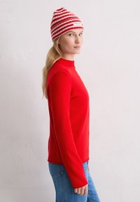 Kobieta z długimi blond włosami, nosząca czerwoną czapkę w paski, czerwoną bluzę z długim rękawem i niebieskie dżinsy, stojąca na boku przy białej ścianie.