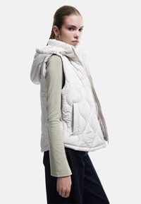 Quiltet hvid ærmeløs vest med hætte, med lynlås foran og sidelommer, båret over en olivengrøn langærmet skjorte. Mørke jeans er synlige.