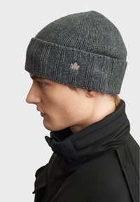 Gorro gris de punto con textura acanalada y un pequeño detalle de una hoja de arce beige en el frente, colocado sobre una chaqueta negra con cuello.