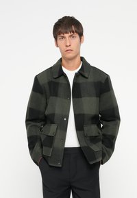 Grüner und schwarzer karierter Wolljacke mit Kragen, Druckknopfverschluss und zwei Vordertaschen. Über einem weißen Hemd getragen.