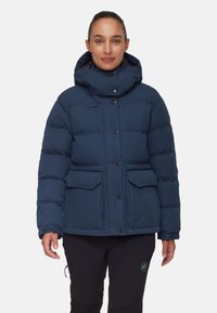 Marineblauwe gewatteerde jas met een hoge kraag, knoopsluiting en twee voorzakken. Beschikt over een capuchon en gewatteerde delen voor isolatie.