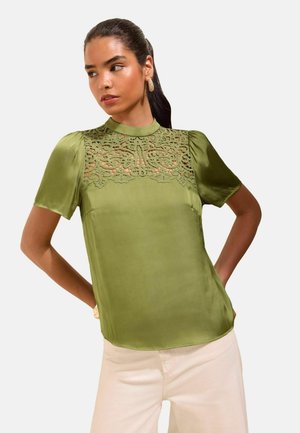 Femme aux cheveux foncés portant un chemisier en satin vert avec un détail en dentelle sur la poitrine et des manches courtes bouffantes, associé à un pantalon clair.