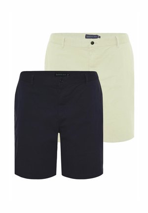 Mand iført en mørkeblå t-shirt, beige shorts og hvide sneakers stående med den ene hånd i lommen mod en ensfarvet baggrund.