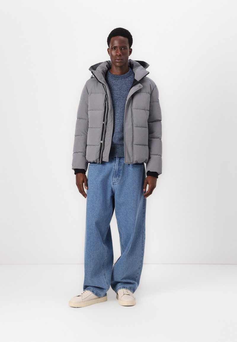 Chaqueta de plumón gris con un diseño acolchado, capucha y cremallera frontal, combinada con jeans holgados de color azul claro y zapatillas blancas.