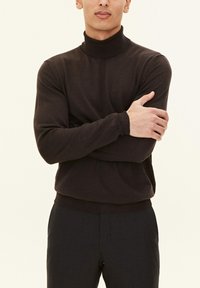 Maglione a dolcevita marrone scuro con un design aderente, maniche lunghe e texture a coste, abbinato a pantaloni grigi.