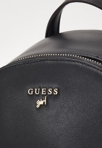 Czarny skórzany plecak o zaokrąglonym kształcie, złotym wykończeniu, z zamkiem błyskawicznym i wyraźnie widocznym logo "GUESS girl".