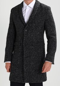 Svart och grå yllekappa med fiskbensmönster, med en krage med skåror, två knappar och sidofickor, med ett enfärgat tyg.