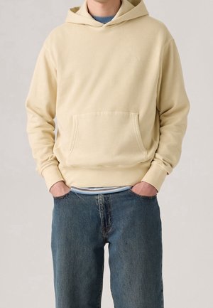 Mand iført en lysebeige hættetrøje med forlommer og blå jeans, hænderne puttet i lommerne, mod en ensfarvet baggrund.