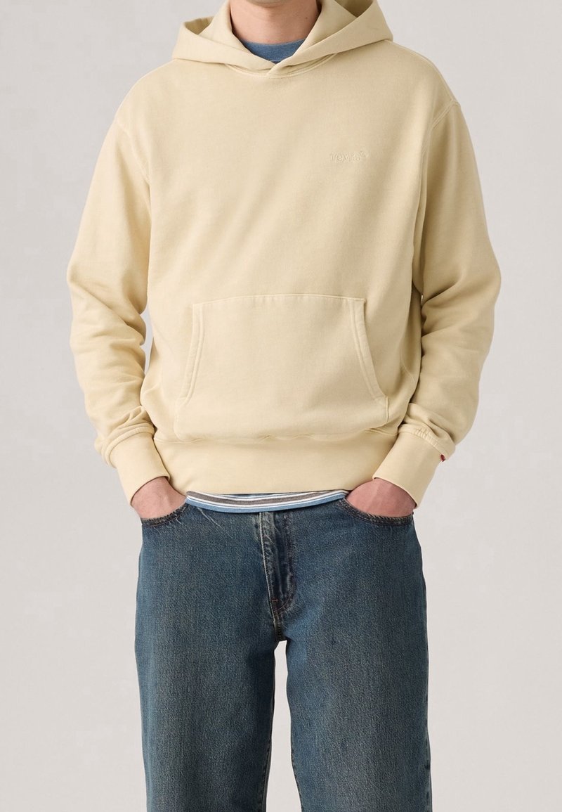 Homme portant un sweat à capuche beige clair avec une poche frontale et un jean bleu, mains dans les poches, sur fond uni.