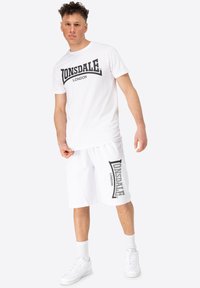 Bílý bavlněný tričko s černým logem "LONSDALE LONDON", spárované s bílými kraťasy se stejným logem po straně. Bílé tenisky outfit dokončují.