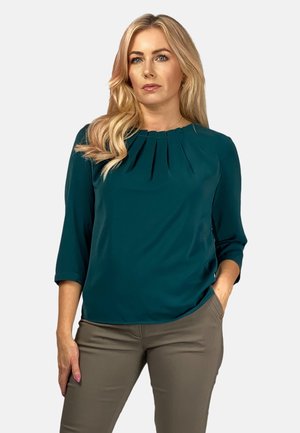 Turkusowa bluzka z rękawami do trzech czwartych, z plisami przy dekolcie. Gładki materiał o luźnym kroju, zestawiona z dopasowanymi beżowymi spodniami.