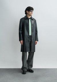 Long manteau en cuir noir avec une fermeture à boutons, associé à un pull vert clair, un pantalon gris à jambes larges, et des chaussures noires.
