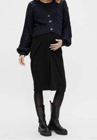 Gravid kvinna i svart klänning, marinblå stickad kofta med knappar, svarta strumpbyxor och svarta stövlar till mitten av vaden, med handen vilande på magen.