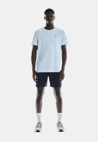 Lichtblauw sportshirt met korte mouwen en een logo, gecombineerd met marineblauwe shorts. Het model draagt witte sokken en grijze sportschoenen.
