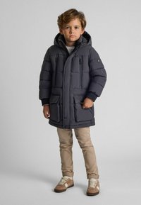 Mørkegrå dunjakke med hætte, lynlåslommer og justerbare manchetter. Båret over beige bukser og brune sneakers. Glat tekstur, firkantet form.