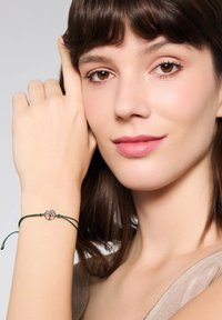 Regulowany zielony sznurkowy bransoletka z okrągłym metalowym zawieszką z motywem drzewa. Modelka prezentuje bransoletkę na nadgarstku, dotykając twarzy.