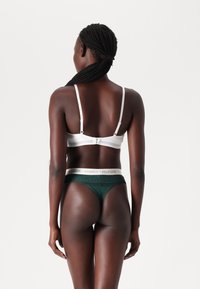 Woman with braided hair wearing a white bra and dark green Tommy Hilfiger underwear, facing away against a plain white background.